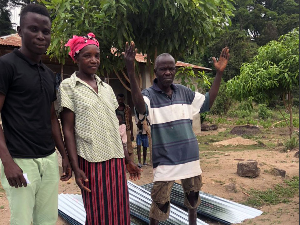 Three members of Mark's African family standing by new roofing with lush greenery behind them. 