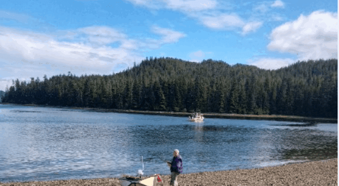 Person by boat on rocky shore, forested lake view.